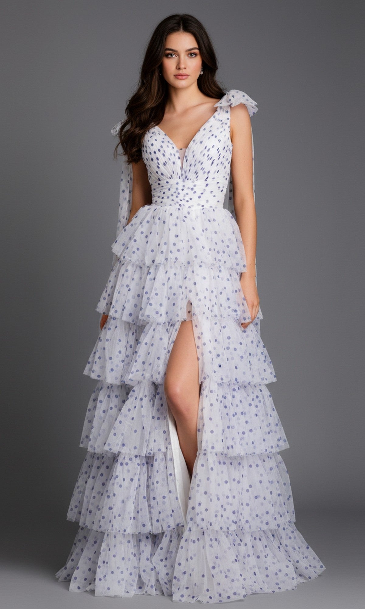 Woman wearing the Jovani 49358 long prom dress featuring white tulle with blue polka dots, deep V-neckline, shoulder ties, and a high slit, standing against a plain gray background.